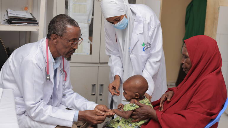 Eine Untersuchung von einem Kind in Somalia - der Arzt hält prüfend und liebevoll die Hand des Mädchens, das auf dem Schoß der mutter sitzt. Die Schwester gibt dem Kind Stabilität.